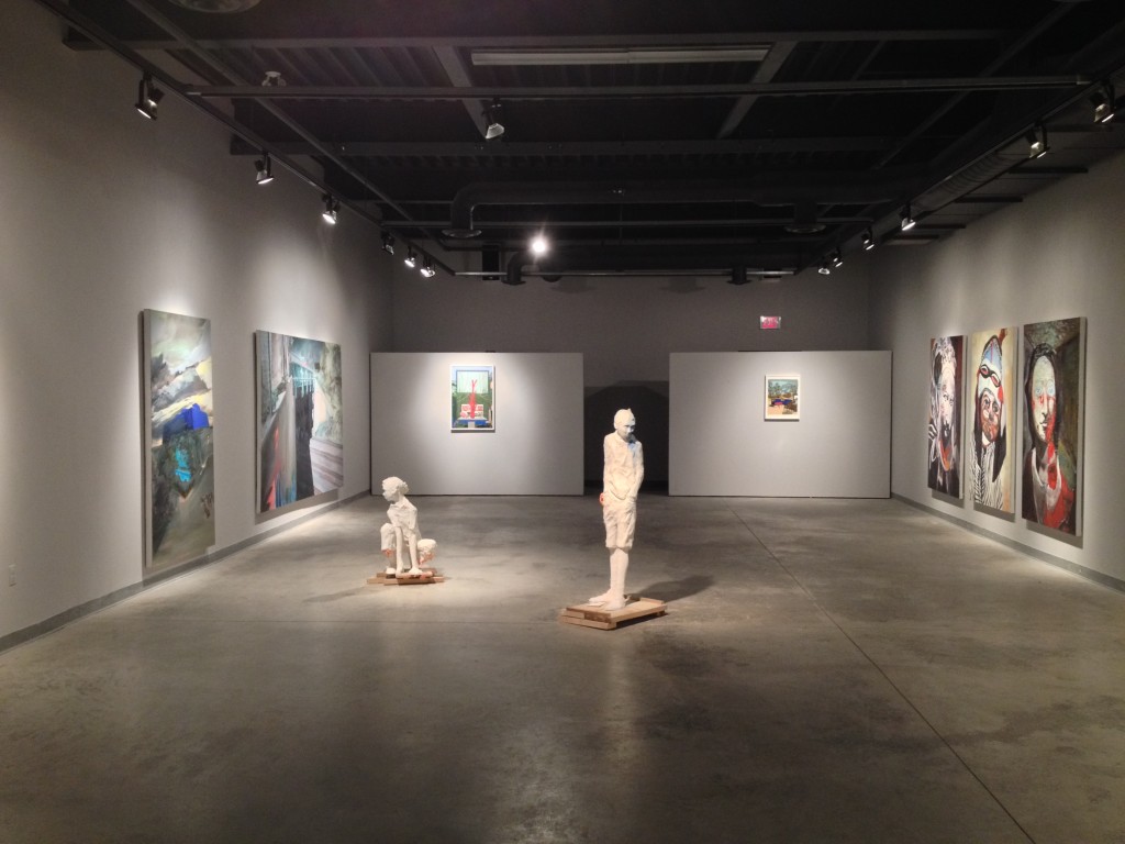 An exhibition of paintings and sculptures in the Campus Gallery. The paintings feature stylized portraits and scenes. The two sculptural works in the centre are two figures, one standing with hands in pocket, and the other crouching down. 