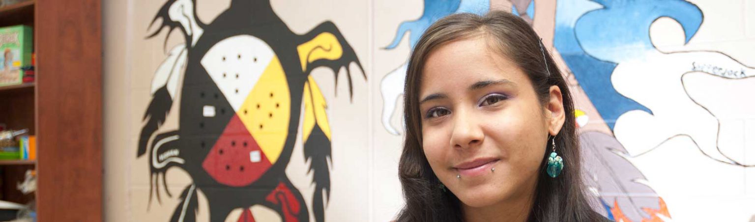 Indigenous Studies student at Georgian College standing in front of a mural featuring an Indigenous medicine wheel