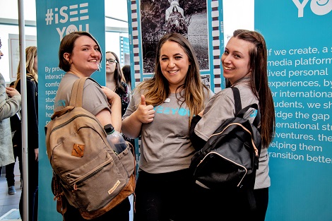 Three females wearing grey, white and teal #iseeyou movement t-shirts, wearing backpacks and standing in front of two pop up banners that showcase their work