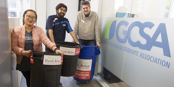 Three people in the GCSA office holding Georgian FoodLocker donation bins