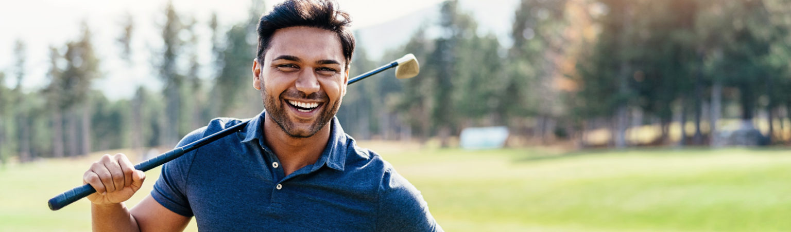 A golf industry management professional wearing a dark blue golf shirt with a golf club over their shoulder, smiling on a golf course
