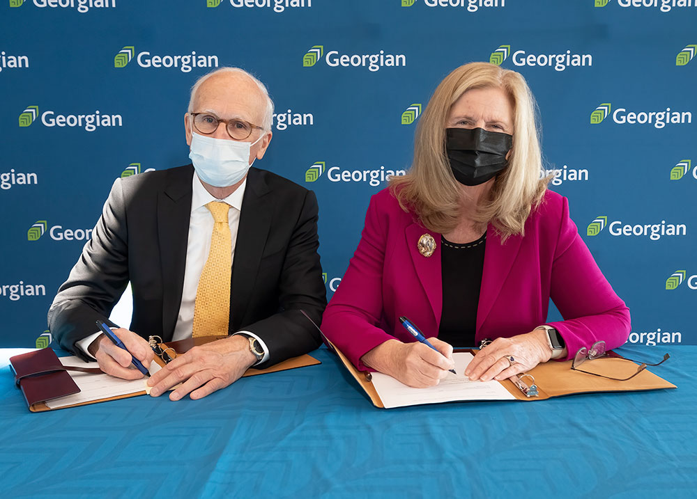 An older white man and woman dressed in business attire sitting at a table signing a document. There is a blue backdrop behind them with the Georgian College logo
