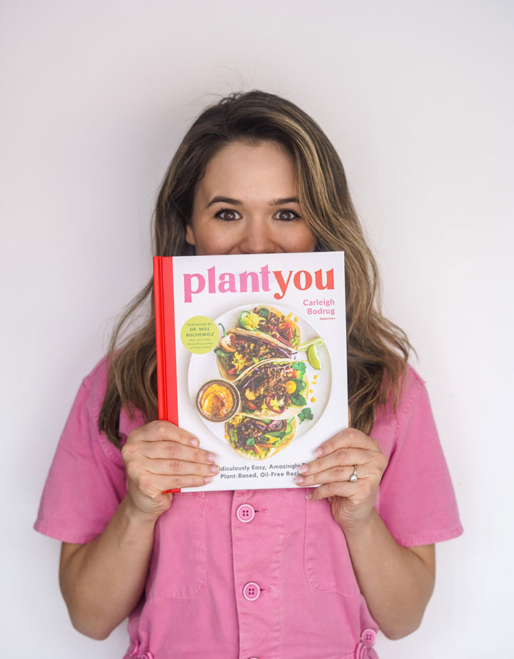 A young woman with long brown hair wearing a pink shirt holding up a book entitled "PlantYou"