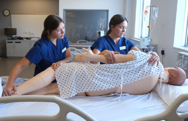BScN students Martha and Anna Koski turning over a human patient simulator on a hospital bed in the Barrie Campus nursing lab