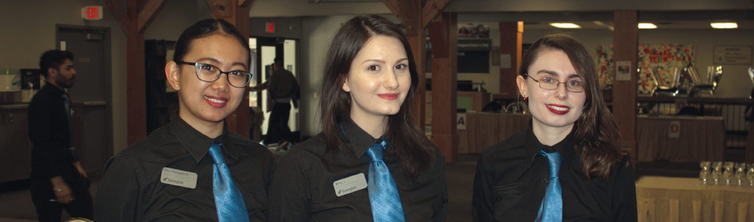 Three Hospitality students wearing black collared shirts, blue silk ties and name tags standing in the Georgian Dining Room