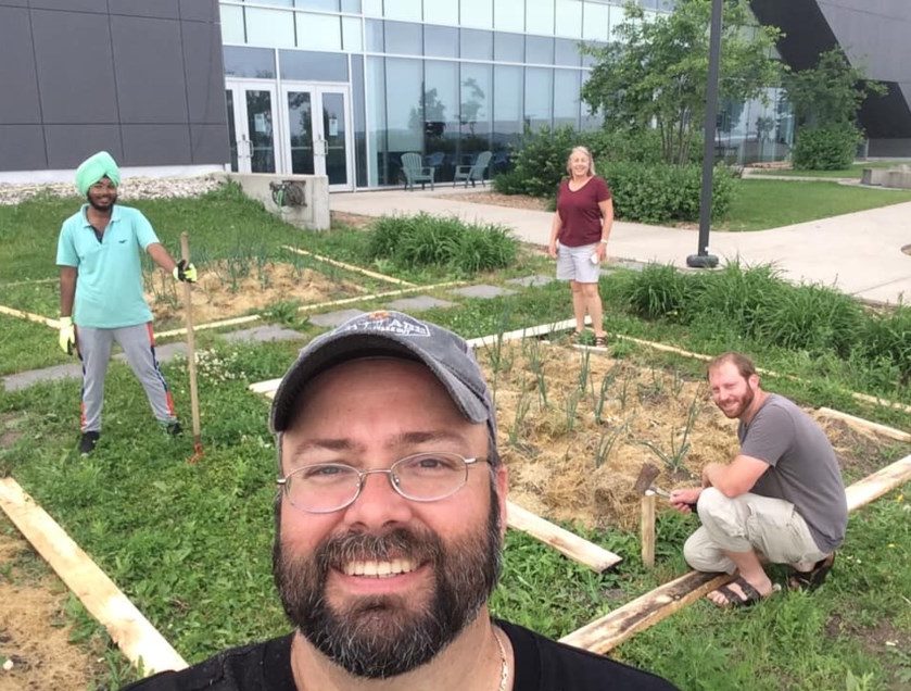 A beared smiling man taking a selfie with three young people standing in the background. They're all in a garden.