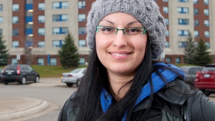 female student with black hair and glasses wearing a hat, jacket and backpack standing in front of the Barrie Campus Residence