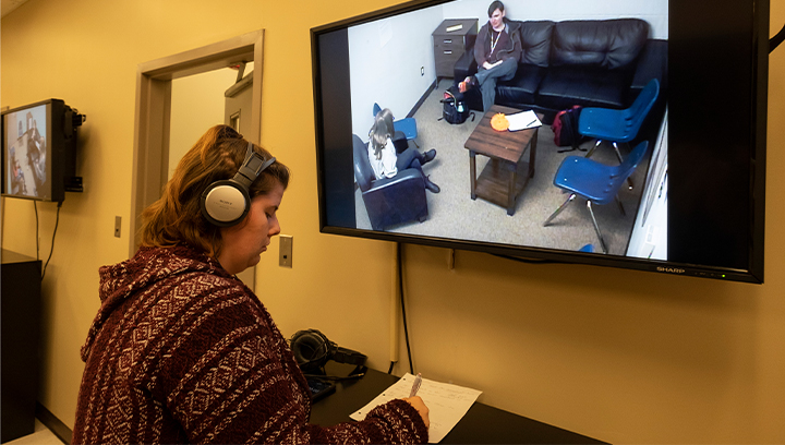 female student wearing a headset and taking notes while watching a counselling session through a TV screen