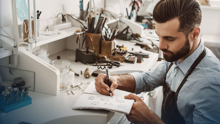 A jewellery designer sitting at a desk in a workshop in a blue collared shirt and brown apron sketching out a design