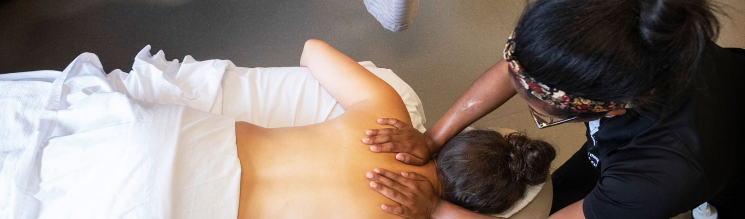 Massage Therapy student in a black collared shirt using her hands to massage the upper back of a laying down patient