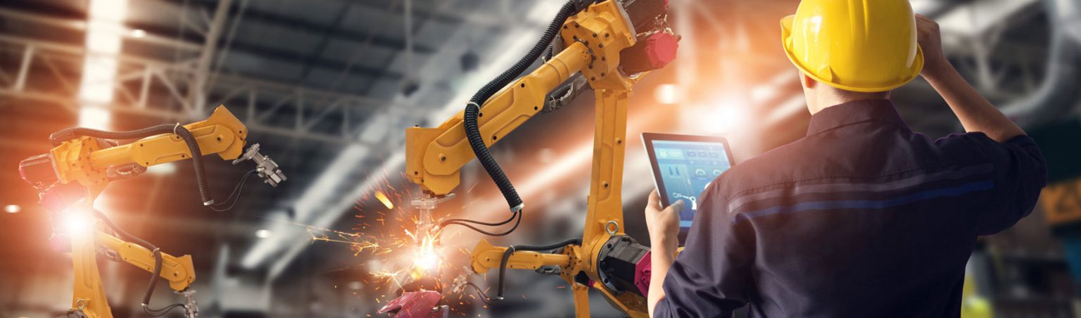 A Mechatronics specialist wearing a yellow construction hard hat holding a tablet and programming a robot