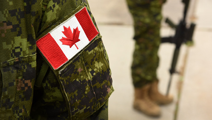 Soldiers in uniform with a Canadian flag patch sewn onto the arm. Solider in background holding an assault rifle gun resting toward the ground.