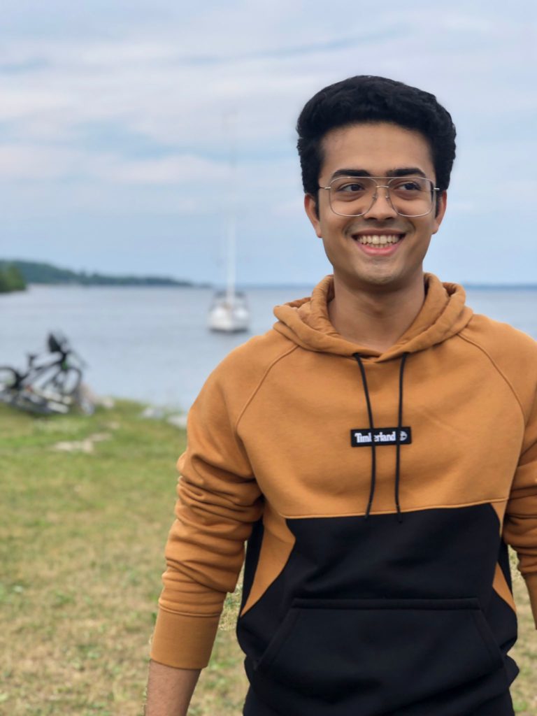 A person wearing a brown sweatshirt and glasses smiles and looks off to the side while standing in front of a lake, with a sailboat in the background.