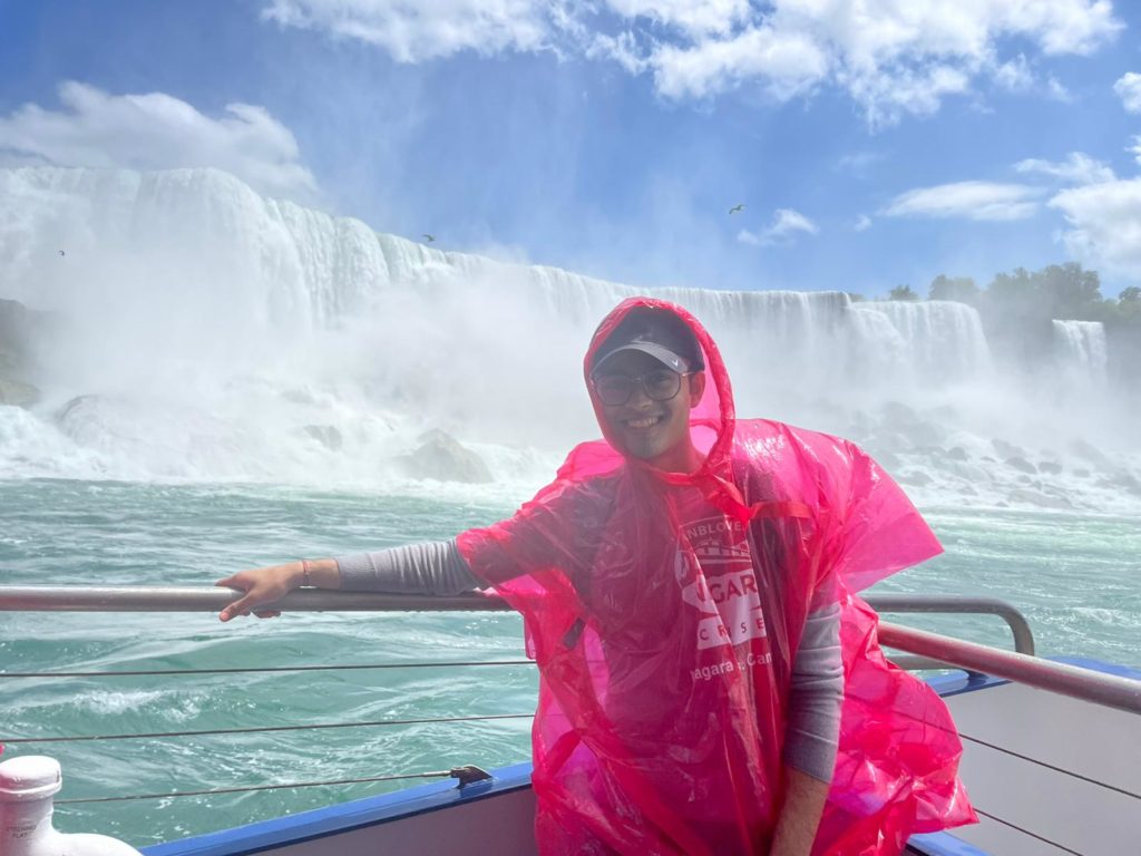 A person with glasses and wearing a red poncho over their clothes, sits on a boat and smiles, with a waterfall in the background.