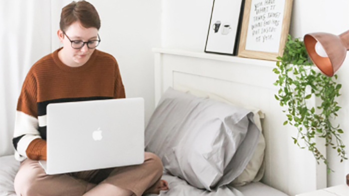 Student sitting cross-legged on a bed while using a Macbook laptop
