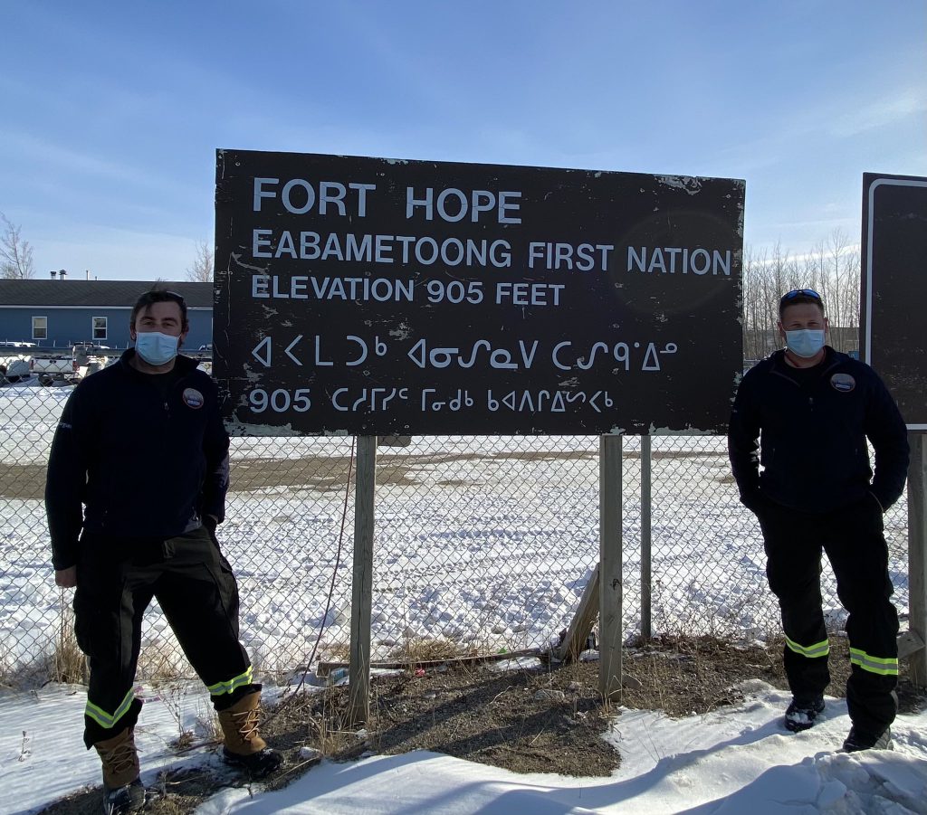 Two people dressed in paramedic uniforms stand next to a sign reading Fort Hope Eabametoong First Nation, elevation 905 feet, in English and an Indigenous language.