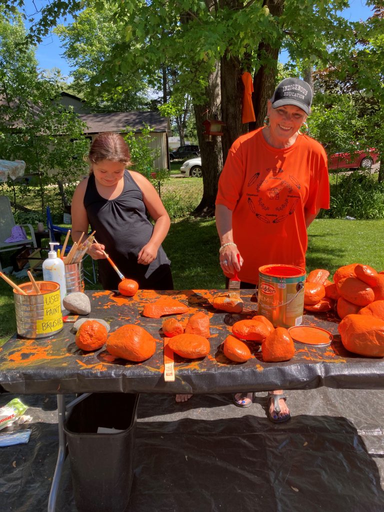 An adult wearing an orange shirt, black shorts, hoop earrings, and black cap reading "Indigenous Education" and a child wearing a black dress stand outside at a table covered in a black tarp and paint rocks orange.
