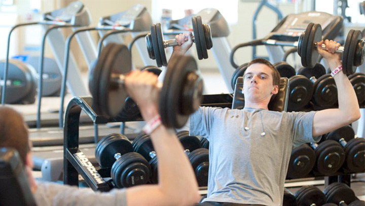 A young man in a grey t-shirt lifting weights inside the Athletics and Fitness Centre at the Georgian College Orillia Campus