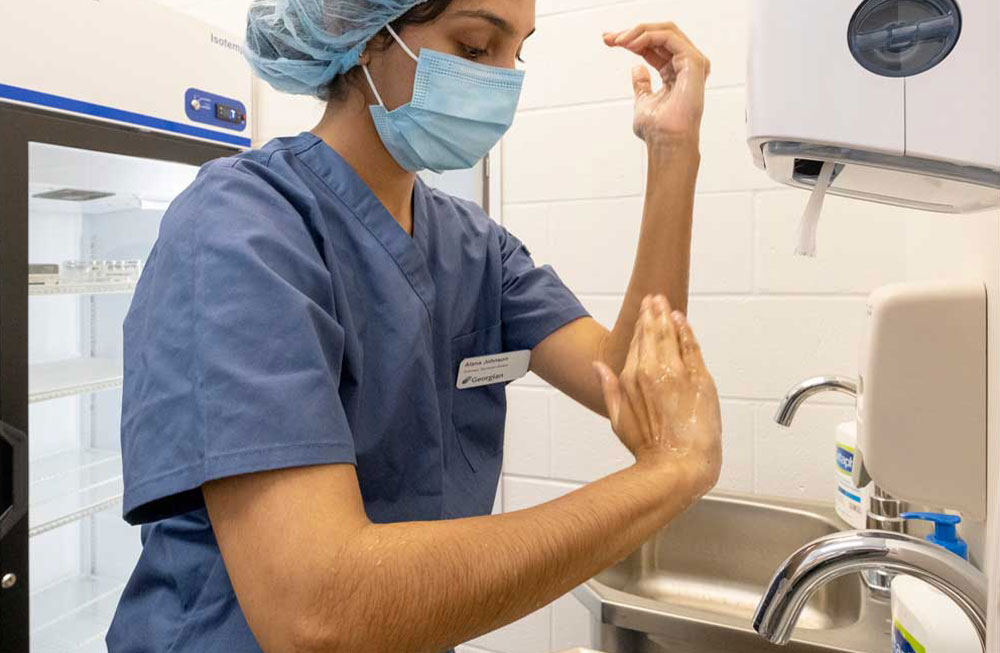 A Georgian College Pharmacy Technician student wearing blue scrubs, a face covering and hairnet washing their hands