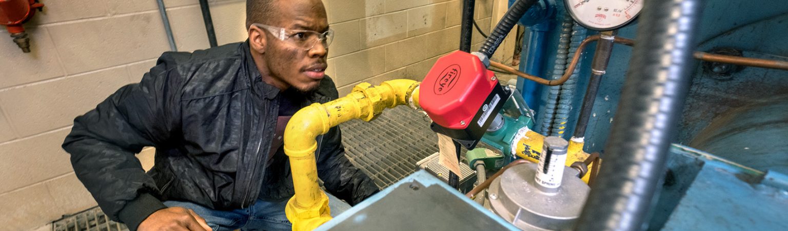 A Plumbing Techniques student wearing safety glasses and crouching while inspecting a piece of equipment