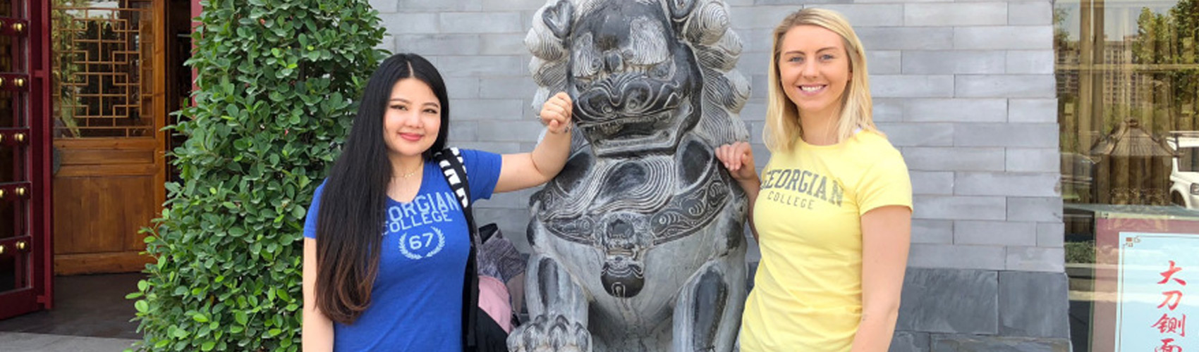Two female students abroad standing next to a stone lion animal statue in China