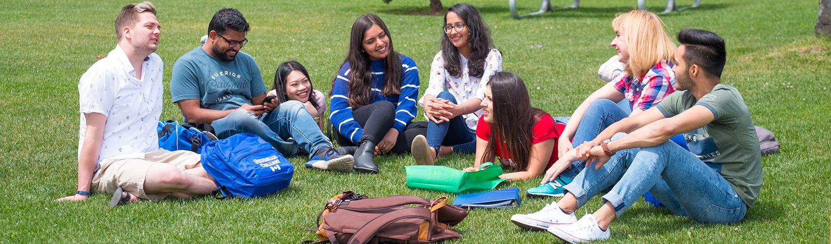 A group of eight international students sitting on the grass with backpacks and binders in between classes at the Barrie Campus