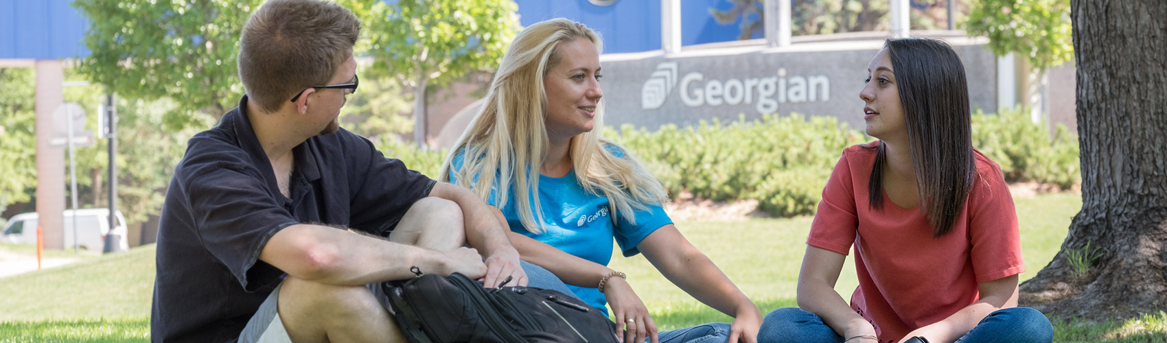 One male and two female students sitting on the grass in front of the main entrance at the Georgian College Barrie Campus