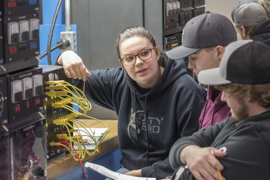 A group of students sitting next to an electrical panel with wires