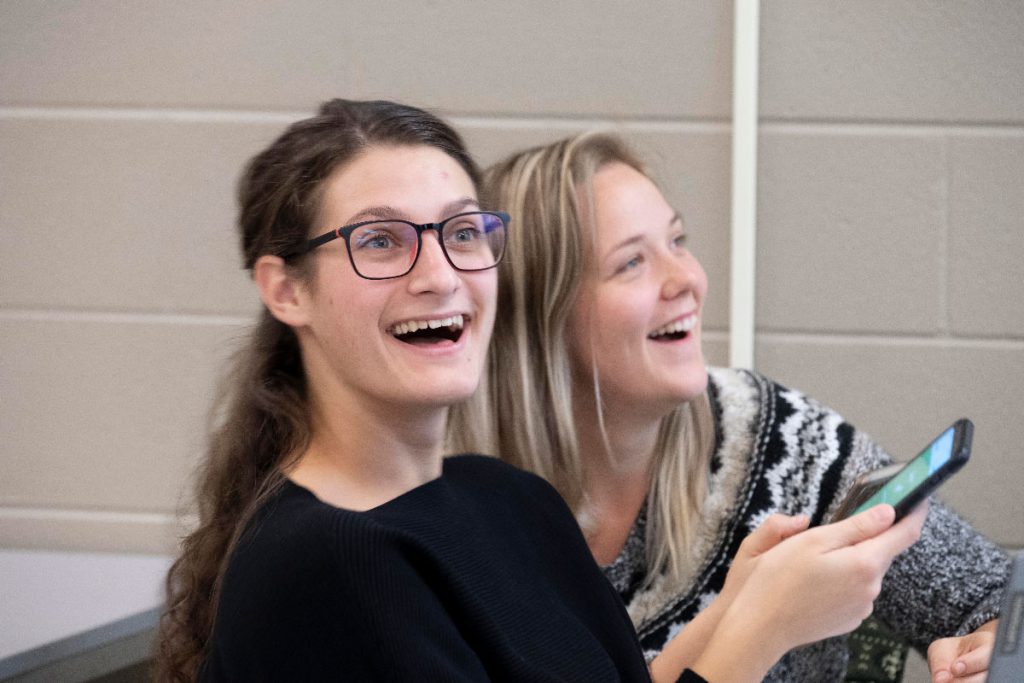 two female students in a classroom smiling and using their cellphones to participate in an in-class exercise