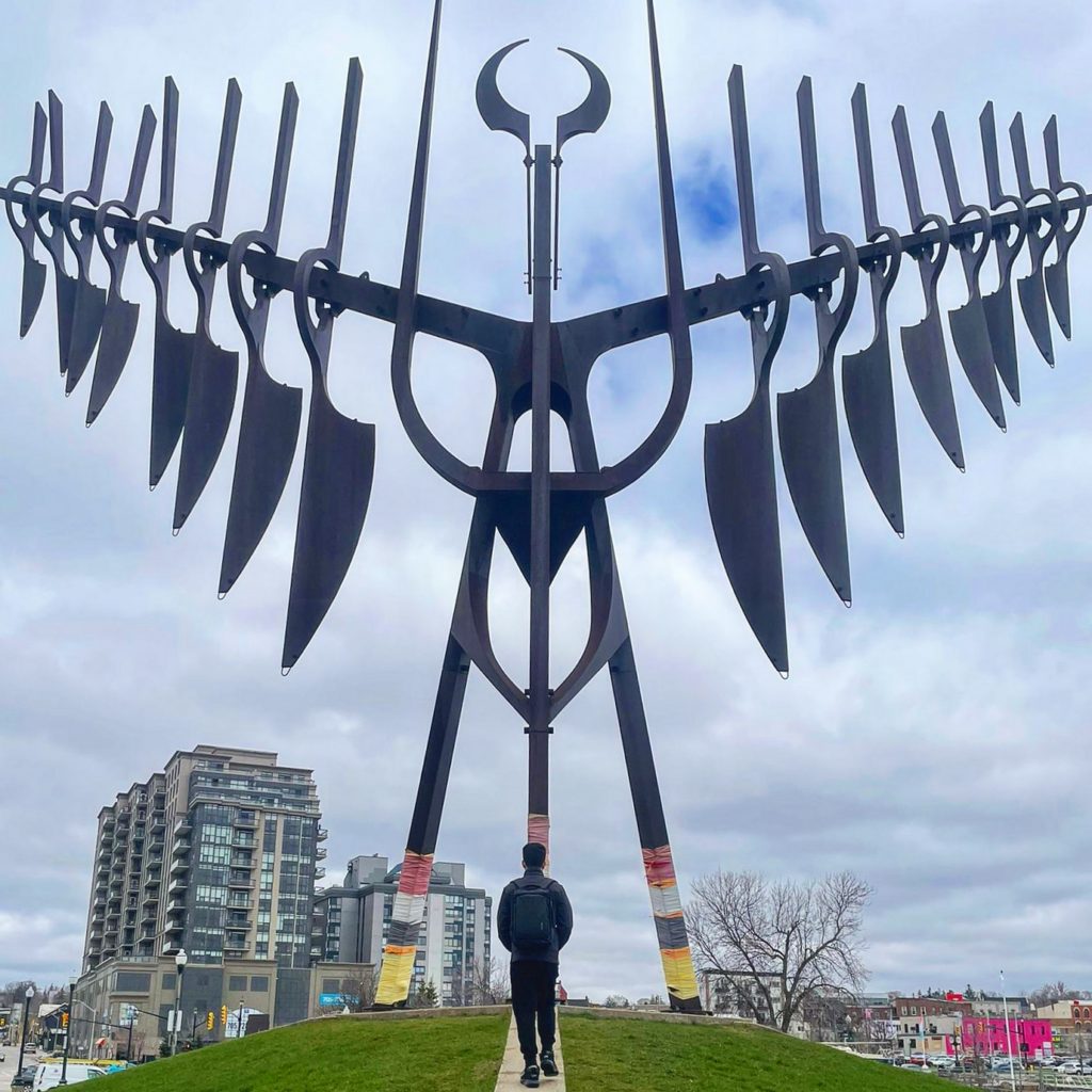Rear view of a person dressed casually with a backpack on waking toward a large Spirit Catcher artpiece in a park, with condo buildings in the background.