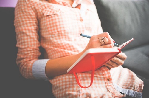 Stock photo from pexels.com of person in orange and white checkered shirt writing in a red journal on a couch; can't see face