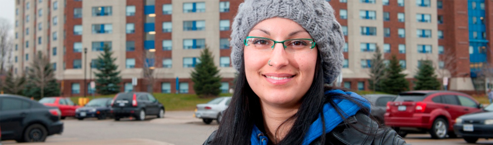 student with black hair, green glasses and a grey knit hat standing in front of Georgian College Barrie Residence