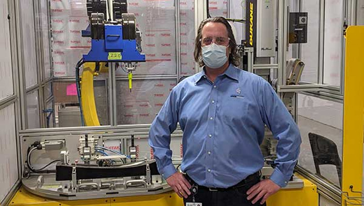 Person in a blue dress shirt, dark jeans, work boots and a mask standing in a factory