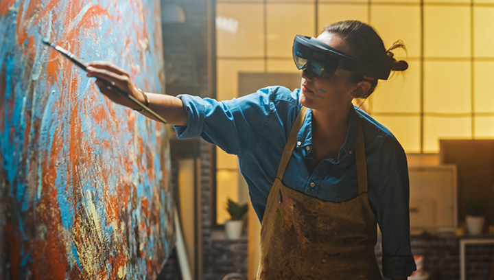 An artist in a blue denim shirt and beige apron wearing a virtual reality headset while painting on a canvas